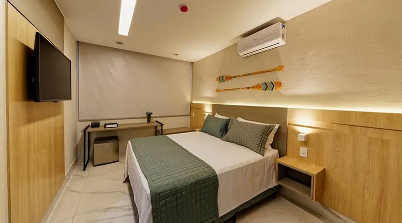 Quarto Standard II: cama de casal queen com colcha verde, criados-mudos, mesa e TV na parede